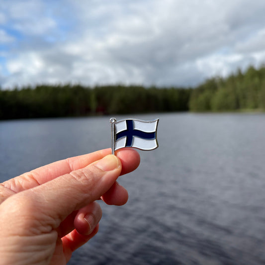 Käsi pitelee Suomen lippu -pinssiä järven ja metsän edessä